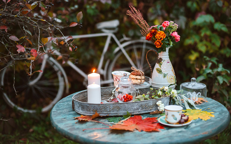 Eine herbstlich dekorierte Tischszene im Freien mit Kerzen, Blumen, Keksen und einer Tasse, mit einem weißen Fahrrad im Hintergrund.