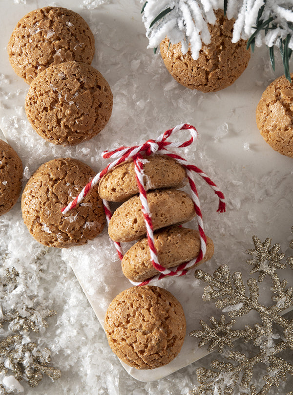 Italienische Amarettini – goldbraune Mandelkekse im Winterstil, mit roter Schnur gebunden, auf Schnee und funkelnden Schneeflocken dekoriert.