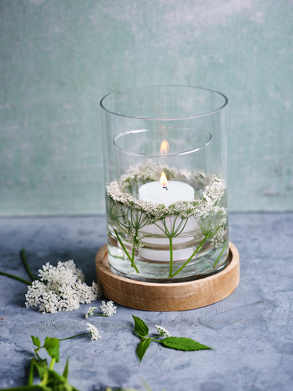 Windlicht mit schwimmender Stumpenkerze in einem doppelten Glasgefäß, dekoriert mit weißen Gierschblüten und Holzuntersetzer – stimmungsvolle DIY-Sommerdeko für Balkon oder Garten.