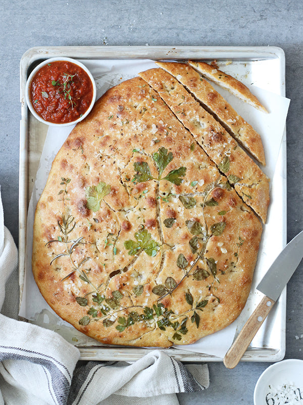 Goldbraun gebackene Focaccia mit frischen Kräutern und grobem Meersalz, angerichtet auf einem Backblech mit Messer und Tomatensugo in einer kleinen Schale.