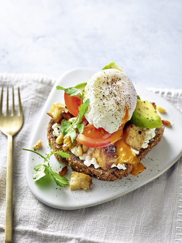 Vollkornbrot belegt mit Hüttenkäse, Avocadospalten, Tomaten, karamellisiertem Honigtofu, Pinienkernen und einem pochierten Ei, garniert mit frischem Koriander – angerichtet auf einem hellen Teller mit goldener Gabel.