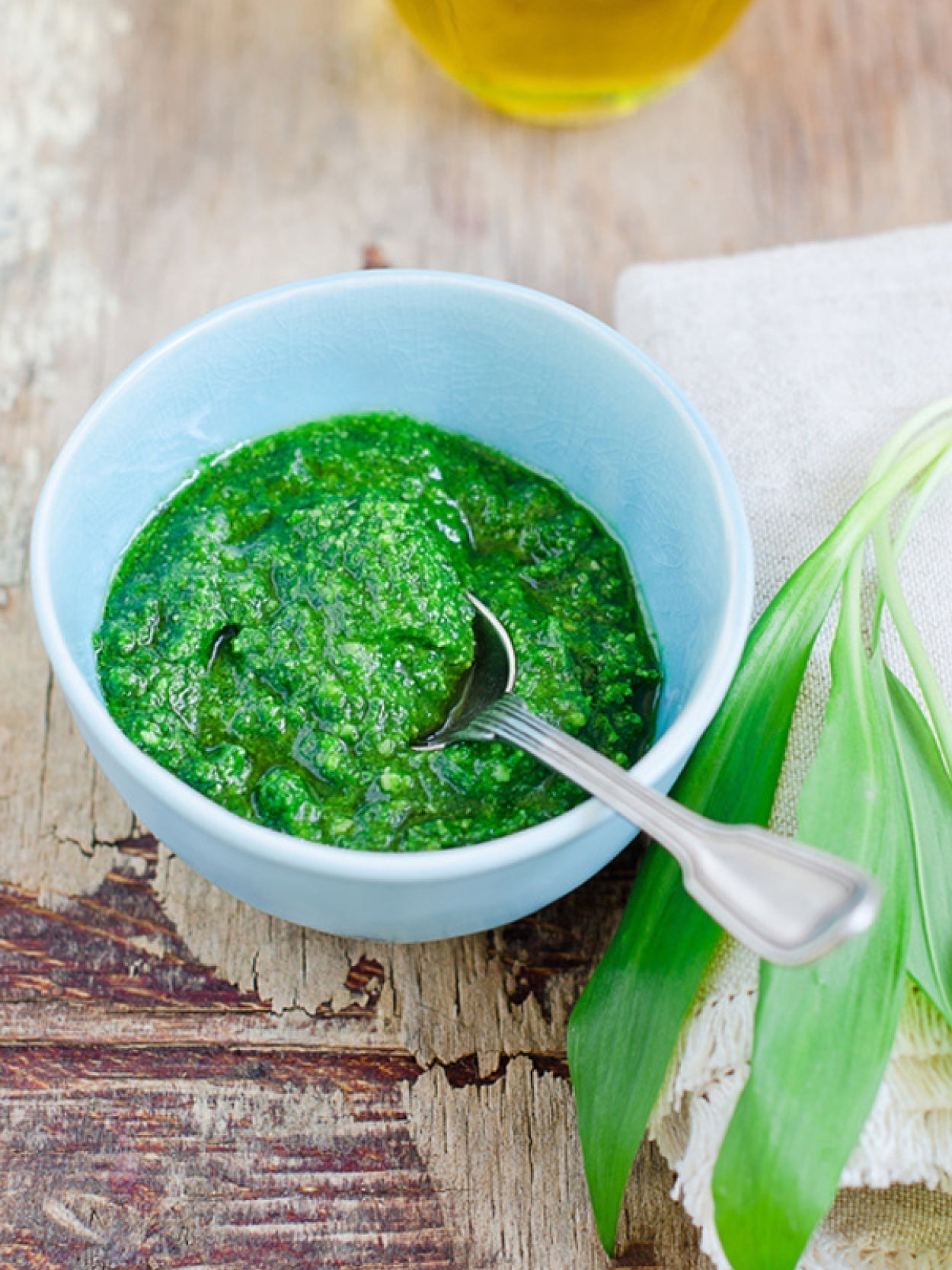Selbstgemachtes Bärlauchpesto in einer hellblauen Schale mit Löffel auf rustikalem Holztisch, daneben frische Bärlauchblätter und Leinentuch.