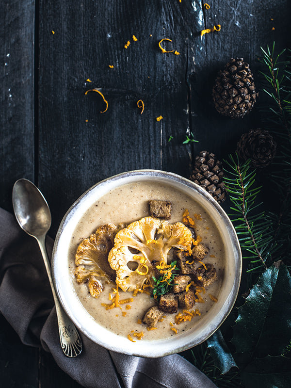Cremige Blumenkohlsuppe in einer Schale, getoppt mit goldbraun gerösteten Blumenkohlscheiben, knusprigen Zimt-Croûtons und Orangen-Petersilien-Gremolata, dekorativ angerichtet auf dunklem rustikalem Holz mit Tannenzweigen und Zapfen.