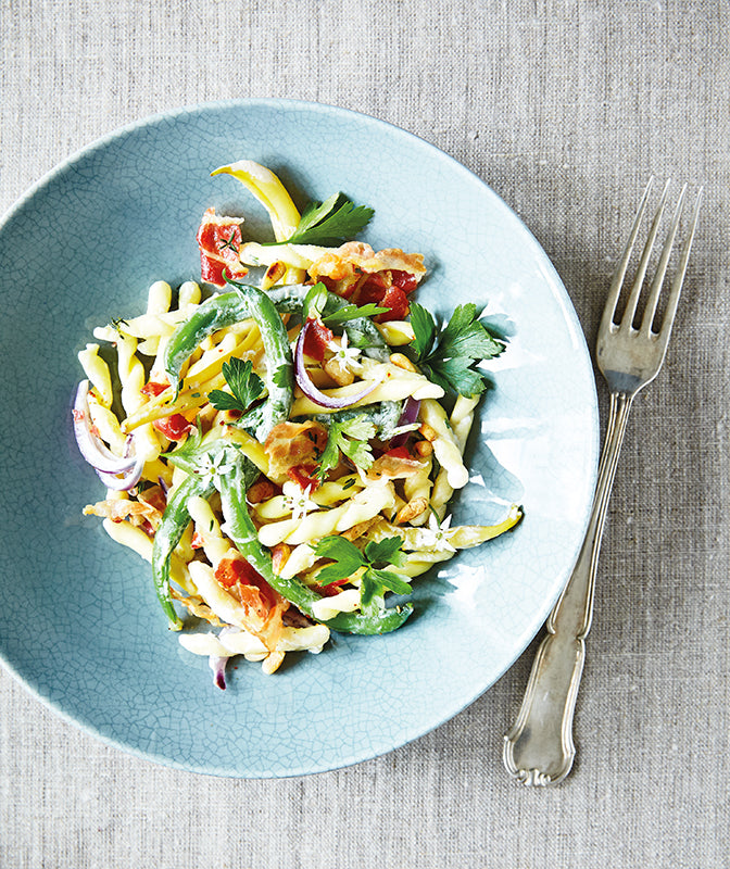 Bohnen-Pasta mit Strozzapreti, grünem Gemüse, knusprigem Pancetta, roten Zwiebeln und Kräutern auf hellblauem Teller – sommerliches Pastarezept mit Gartenbohnen