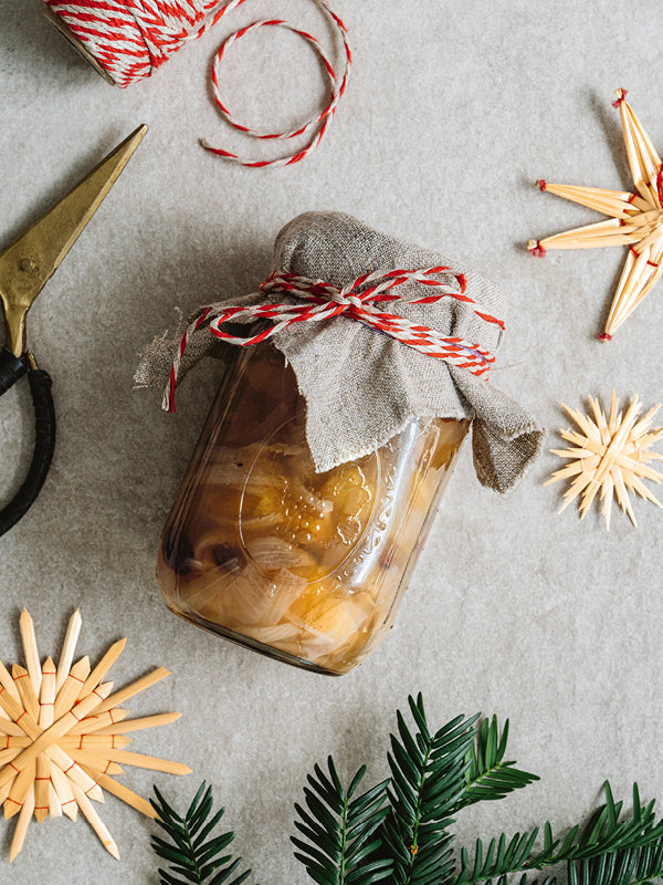 Ein weihnachtlich dekoriertes Einmachglas mit selbstgemachtem Bratapfel-Chutney, mit Stoffhaube und rot-weißer Kordel gebunden, umgeben von Strohsternen, Tannengrün, Schere und Garn.