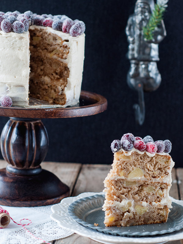 Eine dreistöckige Bratapfel-Torte mit Zimtcreme, gefüllten Apfel-Walnuss-Böden und gezuckerten Cranberrys als Dekoration; ein Stück steht auf einem Teller daneben.