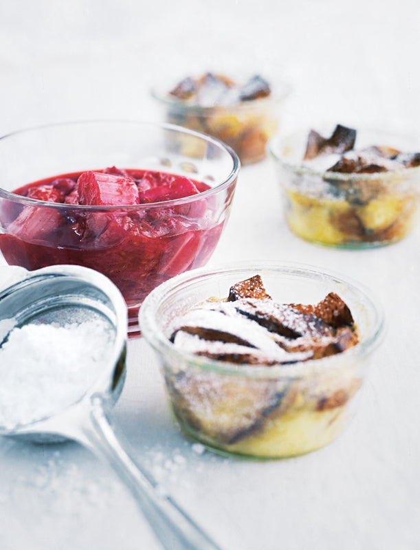 Fruchtiges Rhabarberkompott in einer Glasschale und goldbrauner Brotauflauf in Gläsern mit Puderzucker bestäubt, angerichtet auf hellem Untergrund