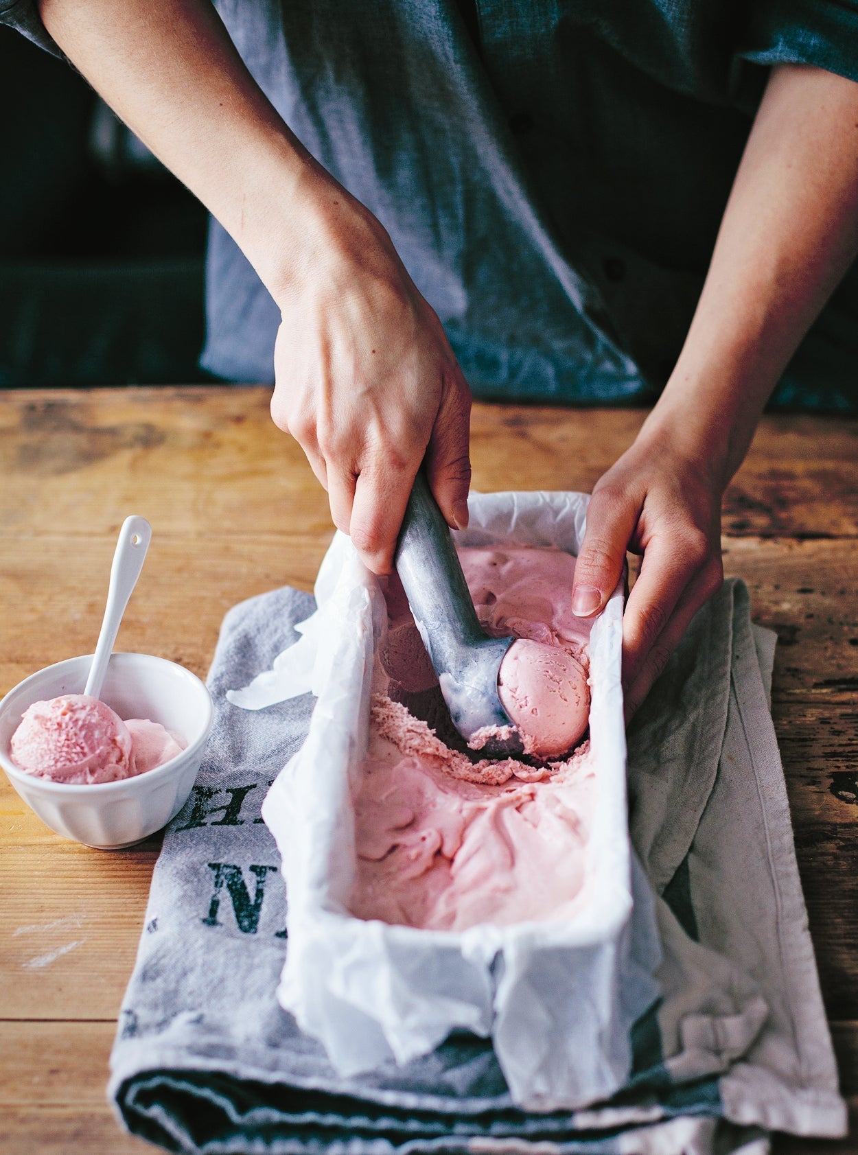 Hausgemachtes Erdbeereis wird aus einer Kastenform geschöpft, daneben eine kleine weiße Schale mit Kugel Eis auf einem Holzbrett mit Stoffserviette.
