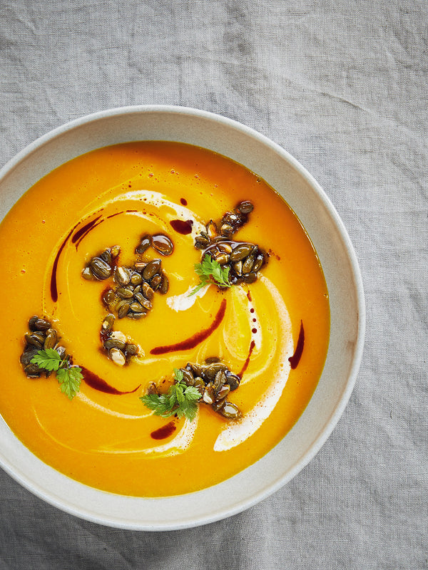 Cremige Kürbissuppe mit Orangen und karamellisierten Kürbiskernen, angerichtet mit Sahne und frischen Kräutern in einem hellen Teller auf Leinentuch.