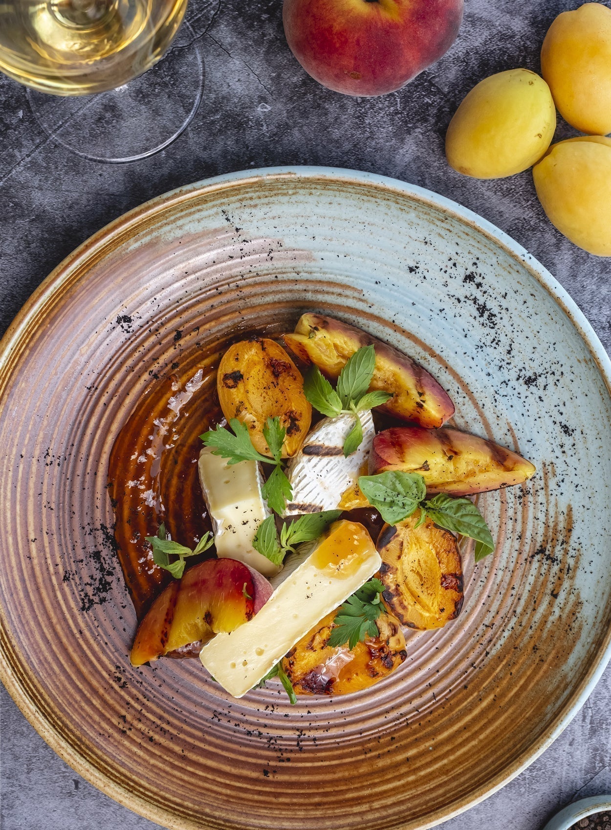 Gegrillte Pfirsiche und Aprikosen mit geschmolzenem Brie und frischen Kräutern, serviert auf rustikalem Keramikteller mit Weißwein im Hintergrund