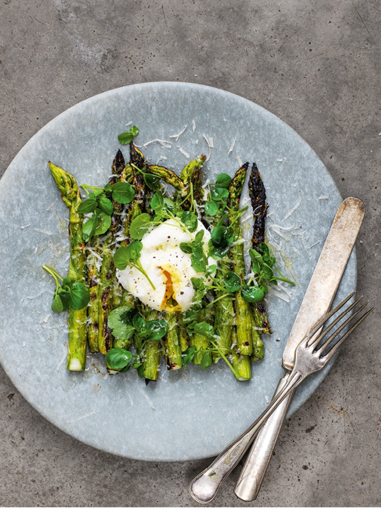Gegrillter grüner Spargel mit pochiertem Ei, frischer Brunnenkresse und geriebenem Parmesan auf hellgrauem Teller – angerichtet mit rustikalem Besteck auf Betonuntergrund.