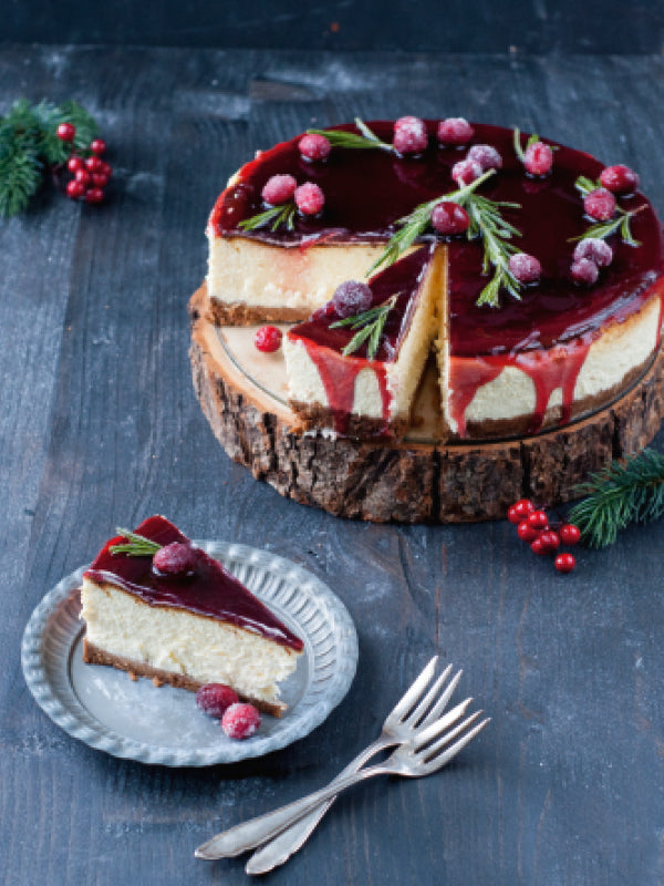 Ein cremiger Cheesecake mit rotem Glühwein-Guss, dekoriert mit Rosmarinzweigen und gezuckerten Cranberrys, auf einer Holzplatte serviert; daneben ein Teller mit einem Stück Kuchen.