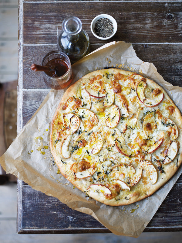 Knusprige Apfel-Taleggio-Tarte auf Backpapier, belegt mit Apfelscheiben, geschmolzenem Käse, Salbei und Olivenöl, auf einem rustikalen Holztisch.