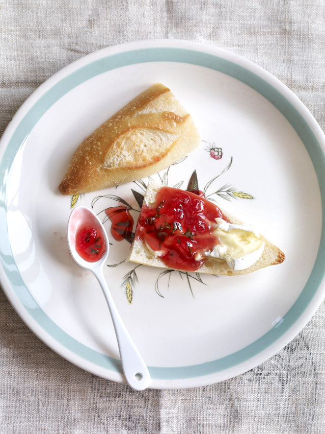 Baguettescheibe mit Brie und Himbeergelee mit Zitronenmelisse auf einem weißen Teller mit floralem Dekor, daneben ein Löffel mit Gelee – sommerlicher Brotaufstrich liebevoll angerichtet.