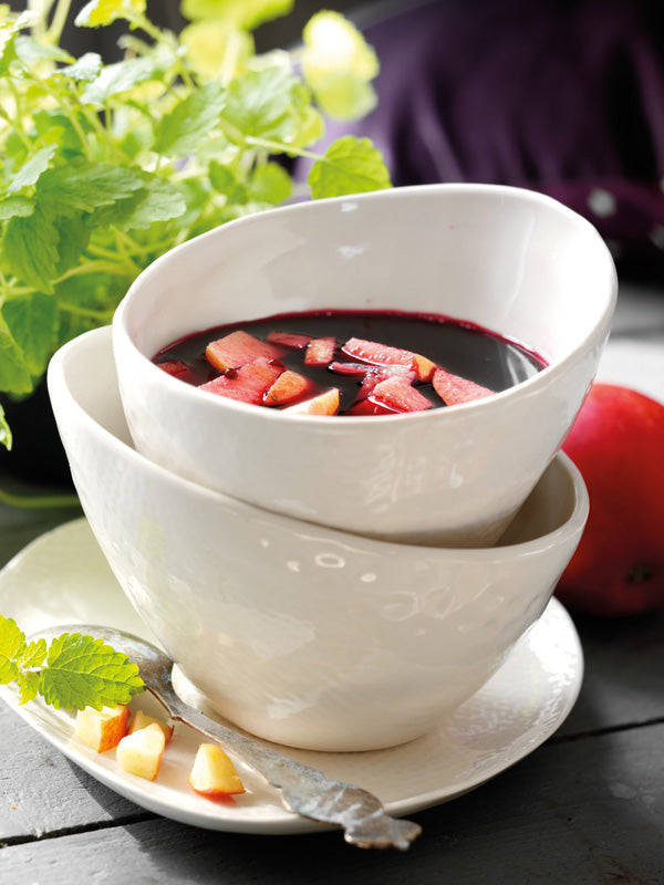 Holunderbeersuppe mit Apfelstückchen, serviert in einer weißen Schale auf einem Unterteller mit Löffel, daneben frische Minze und Apfelstückchen.