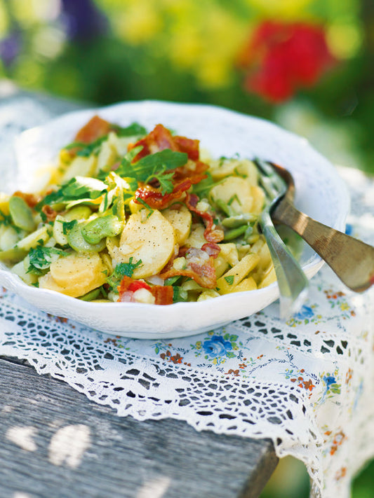 Sommerlicher Kartoffelsalat mit dicken Bohnen, frischem Rucola und knusprigem Speck in einer weißen Porzellanschale, serviert auf einem Holztisch mit nostalgischer Spitzendecke im Garten.