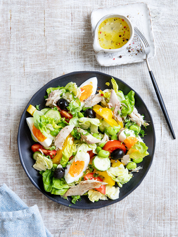 Bunter Nizzasalat mit hartgekochten Eiern, geräucherter Makrele, Bohnen, Oliven und frischem Gemüse auf einem schwarzen Teller – daneben ein kleines Schälchen Zitronen-Dressing auf rustikalem Holztisch.