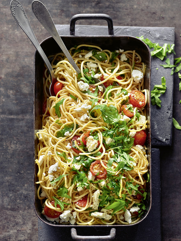 Auflaufform mit Ofen-Spaghetti, Kirschtomaten, Feta und frischem Rucola, serviert mit zwei Löffeln.