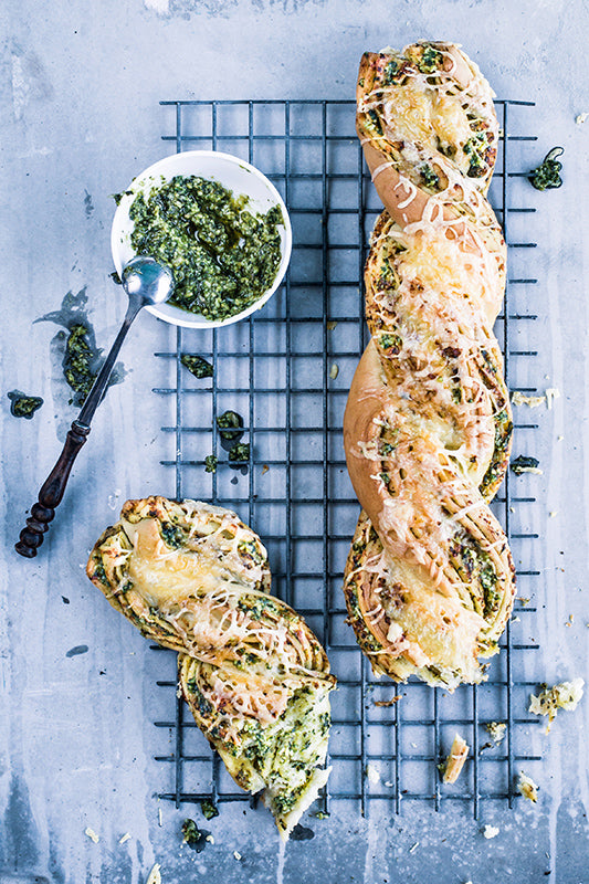 Frisch gebackenes Pestobrot mit geschmolzenem Käse auf einem Kuchengitter, daneben eine kleine Schale Basilikumpesto mit Löffel.