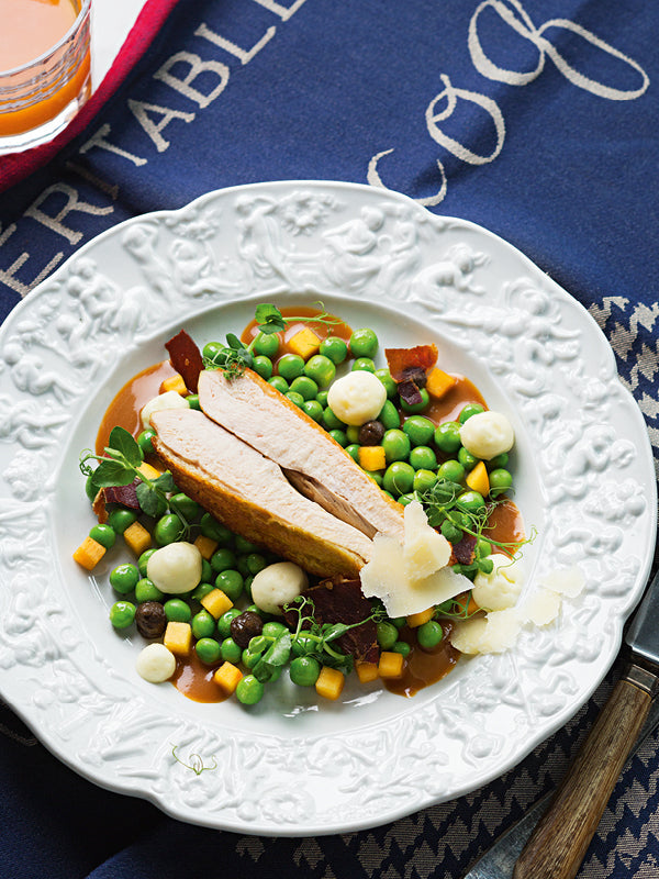 Bresse-Poularde auf Erbsen-Pfirsich-Gemüse mit luftiger Nussbutterschaum-Deko, Speckchips und cremigem Pilzpüree auf edlem Porzellanteller – angerichtet auf dunkelblauem Stoff mit Schriftzug.