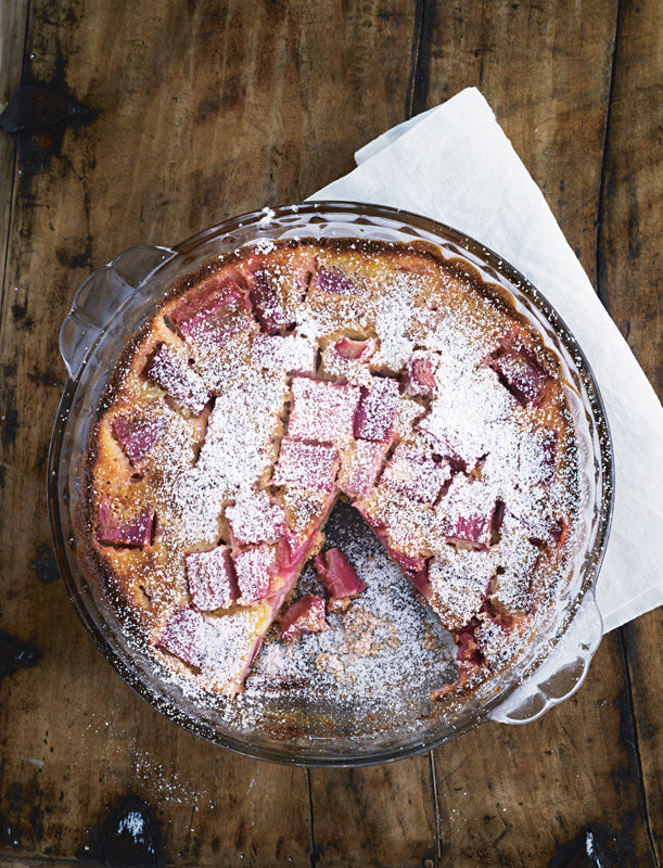 Hausgemachter Rhabarberkuchen mit Puderzucker auf Holzuntergrund, angeschnitten in einer runden Glasbackform