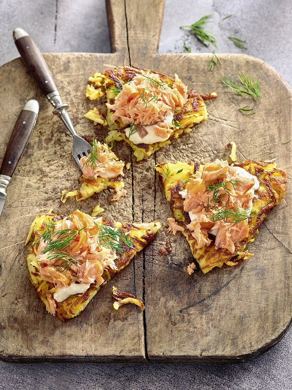 Knusprige Rösti-Stücke mit Sahnemeerrettich, zerpflücktem Stremellachs und frischem Dill auf einem rustikalen Holzbrett.