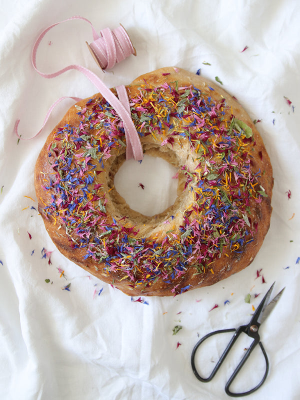 Rundes, goldbraun gebackenes Blütenbrot mit bunten, essbaren Blüten, dekorativ angerichtet auf einem weißen Tuch mit Schleifenband und Schere.