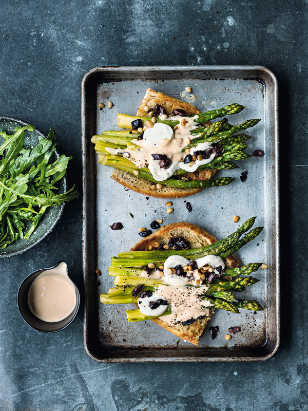 Zwei knusprige Spargelsandwiches mit grünem Spargel, Ziegenkäse, Oliven und Pinienkernen auf einem Backblech, daneben Rucola-Salat und Sauce.