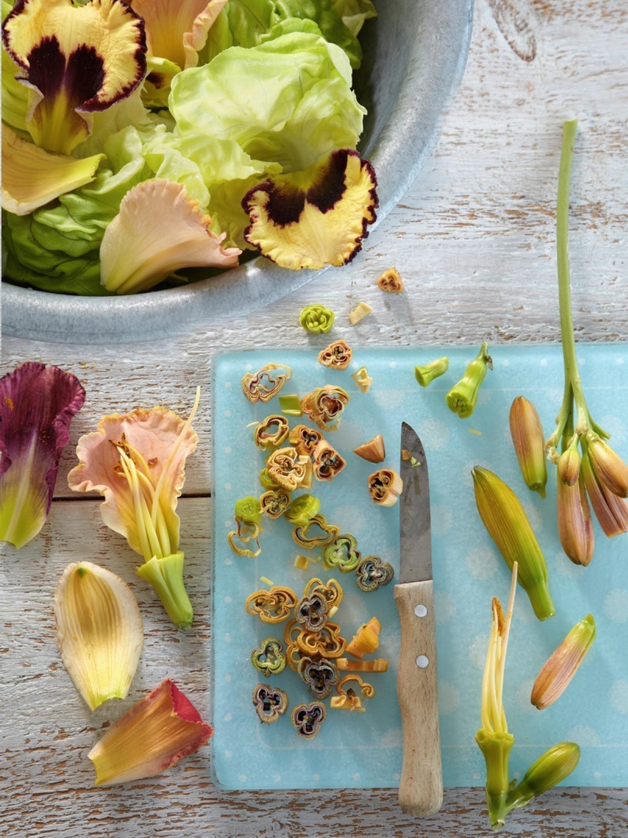 Geschnittene Taglilienknospen auf einem Schneidebrett mit Messer, daneben frische Blattsalate und geöffnete Blüten – Zutaten für einen kreativen Blütensalat.