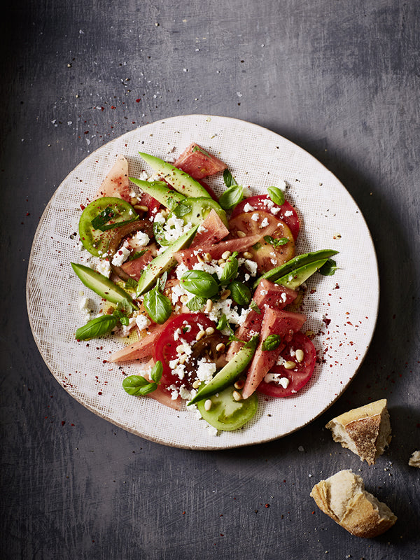 Bunter Tomaten-Melonen-Salat mit Avocado, Feta, Basilikum und Pinienkernen auf heller Keramikplatte