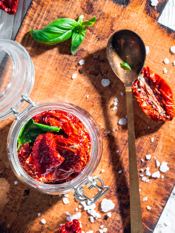 Confierte Kirschtomaten mit frischem Basilikum in einem Einmachglas – daneben getrocknete Tomaten, Knoblauch und Salz auf einem Holzbrett.