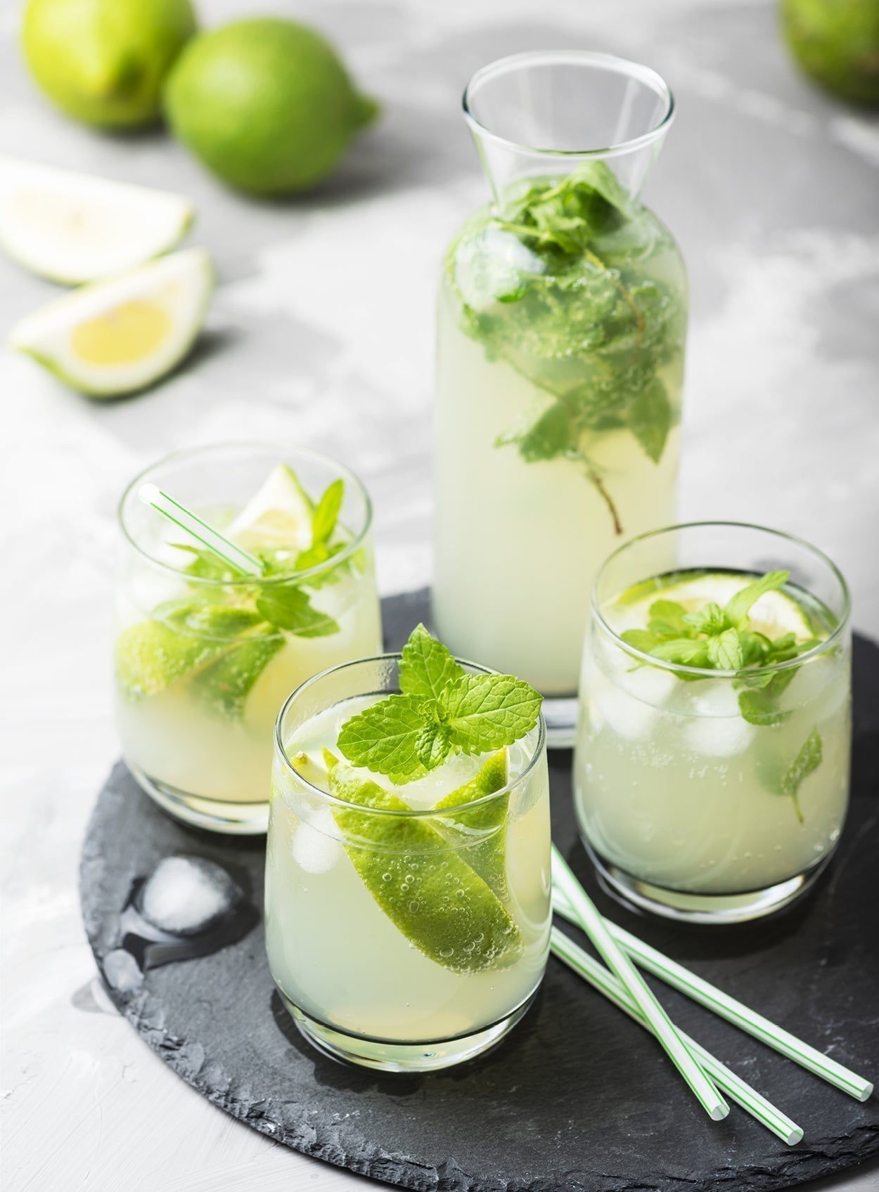 Drei Gläser Virgin Mojito mit frischer Minze, Limettenscheiben und Eiswürfeln, serviert auf einer runden Schieferplatte.