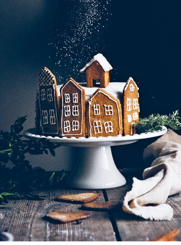 Eine festliche Weihnachtstorte auf einer weißen Tortenplatte, umrahmt von dekorativen Pfefferkuchenhäusern und leicht mit Puderzucker bestäubt, davor Zweige und Gebäckstücke.