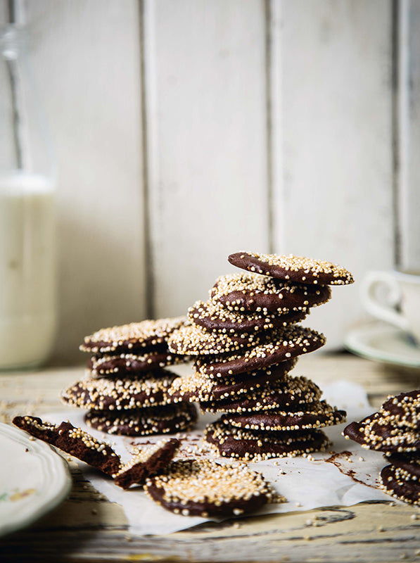 Zarte, knusprige Schokoladenblätter mit Amarant-Pops gestapelt auf Backpapier – dekorativ angerichtet mit Milchflasche und Porzellantasse im Hintergrund.