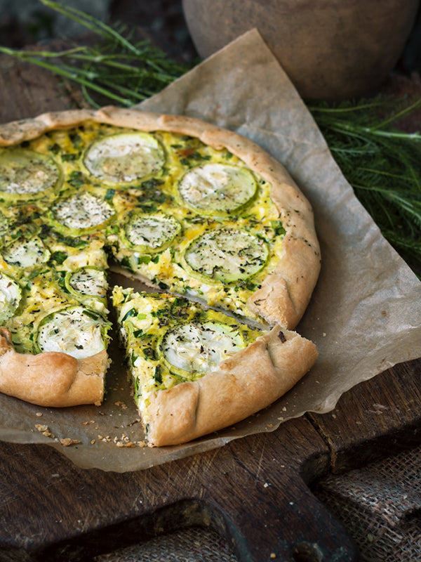 Herzhafte Zucchini-Galette mit Ziegenkäse auf Backpapier – goldbraun gebacken, mit frischen Kräutern bestreut und auf rustikalem Holzbrett serviert.