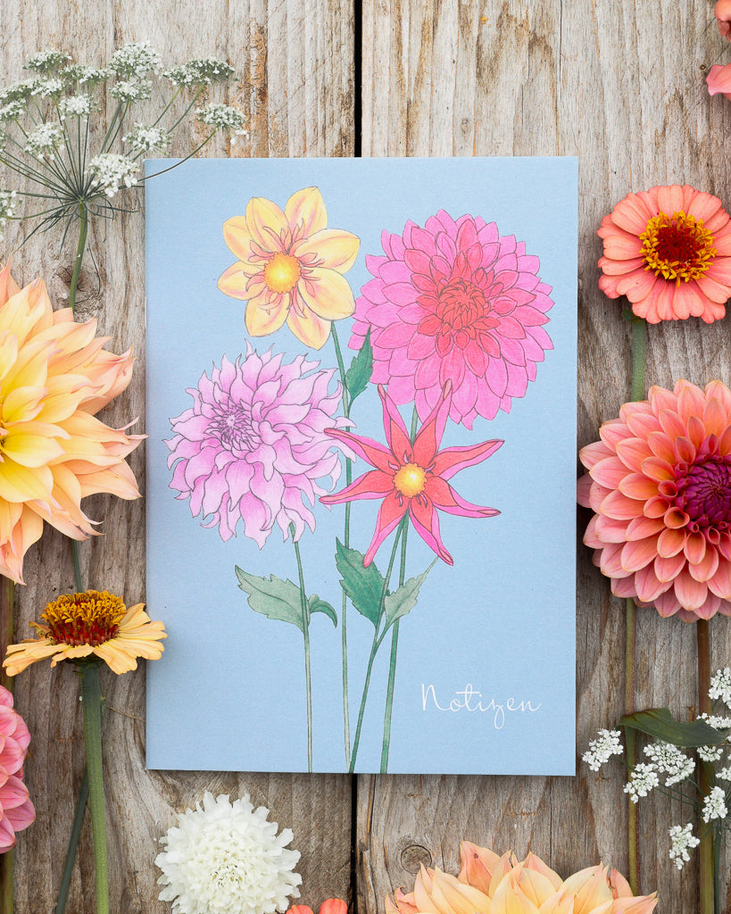 Detailaufnahme des A5 Notizhefts Dahlien – Gartenzauber Papeterie, liegend auf Holztisch mit Dahlienblüten im Hintergrund.