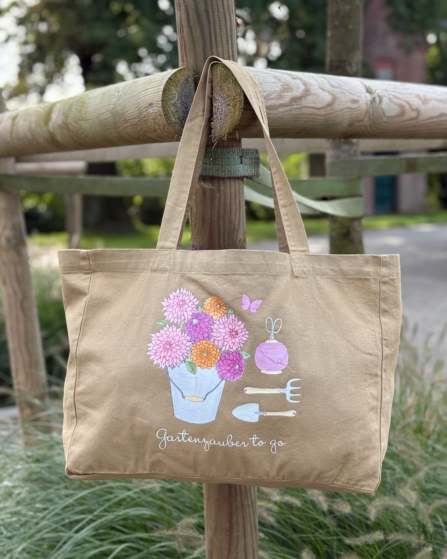„Baumwoll-Shopper ‚Gartenzauber to go‘ im Washed-Look, hängend an einem Holzzaun, bedruckt mit handgezeichneten Blumen- und Gartenmotiven.“