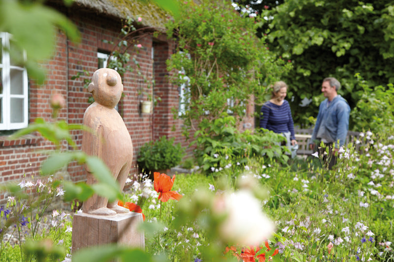 Eine geschnitzte Holzfigur im Staudengarten vor einem alten Bauernhaus.