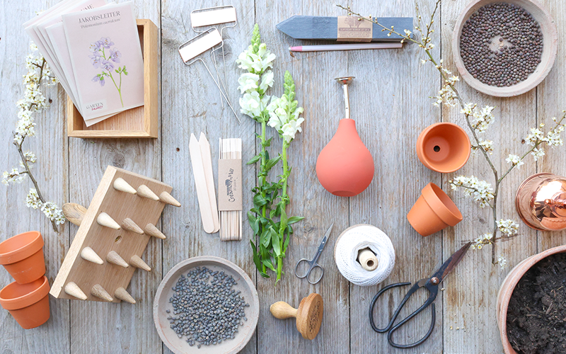 verschiedene Produkte aus der Gartenzauber Kollektion für die Aussaat von Blumen, Saatgutttüten, Löwenmäulchen und Frühlingszweige auf einem Holzbrett.