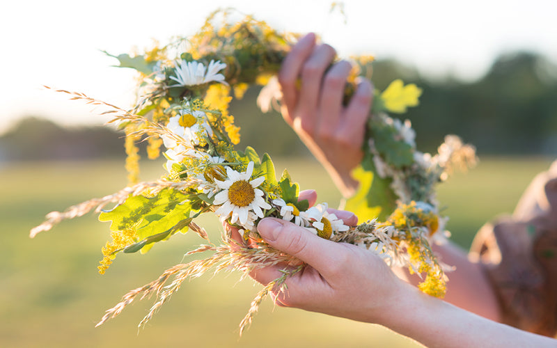 Hände halten einen frisch gebundenen Blumenkranz aus Gänseblümchen, Gräsern und grünem Laub im warmen Sonnenlicht. Im Hintergrund ist eine grüne Wiese unscharf zu erkennen.