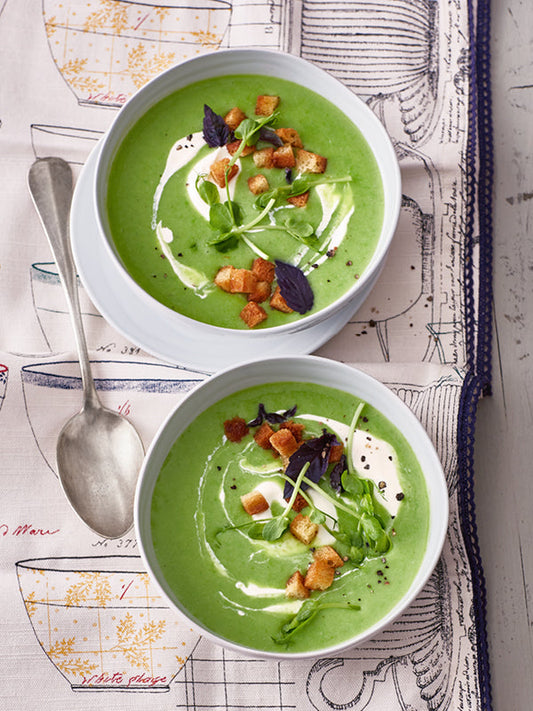 Zwei Schalen grüne Erbsensuppe, garniert mit Crème fraîche, knusprigen Croûtons, Erbsensprossen und Basilikum, serviert auf einem dekorativen Tuch mit Löffel.