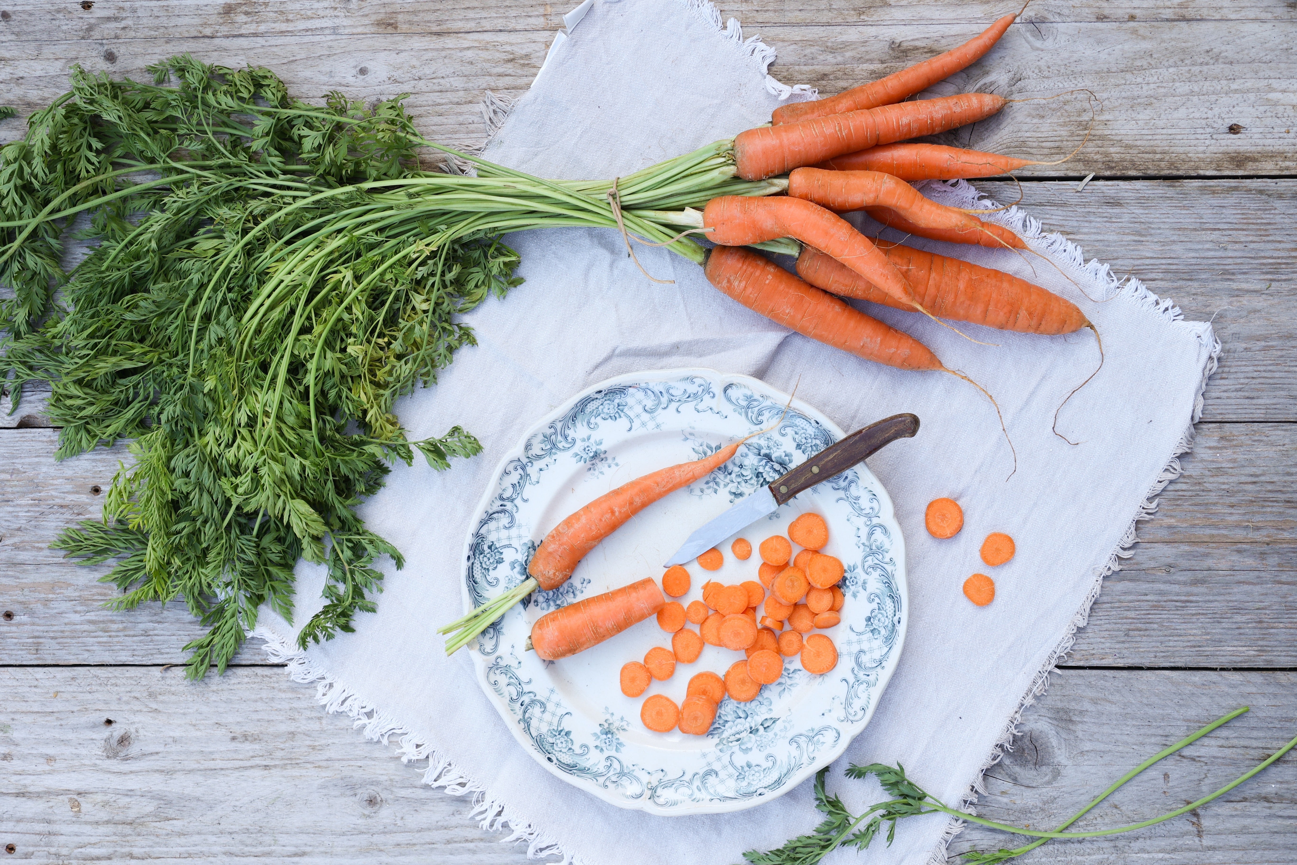 Frisch geerntete Möhren mit grünem Blattwerk auf einem Holztisch, daneben ein Teller mit geschnittenen Möhrenscheiben und einem Küchenmesser – ideal für Garten- und Erntethemen.
