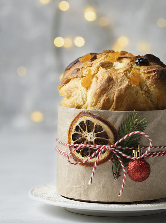 Klassischer Panettone mit Rosinen und kandierten Früchten – in Papier gehüllt, mit getrockneter Orange, Tannenzweig und roter Schleife festlich dekoriert.
