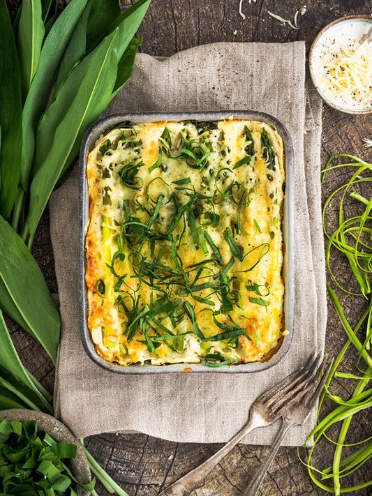 Bärlauch-Spargel-Lasagne in einer Auflaufform, mit goldbrauner Käsekruste und frischen Bärlauchstreifen garniert, serviert auf einem rustikalen Holztisch.