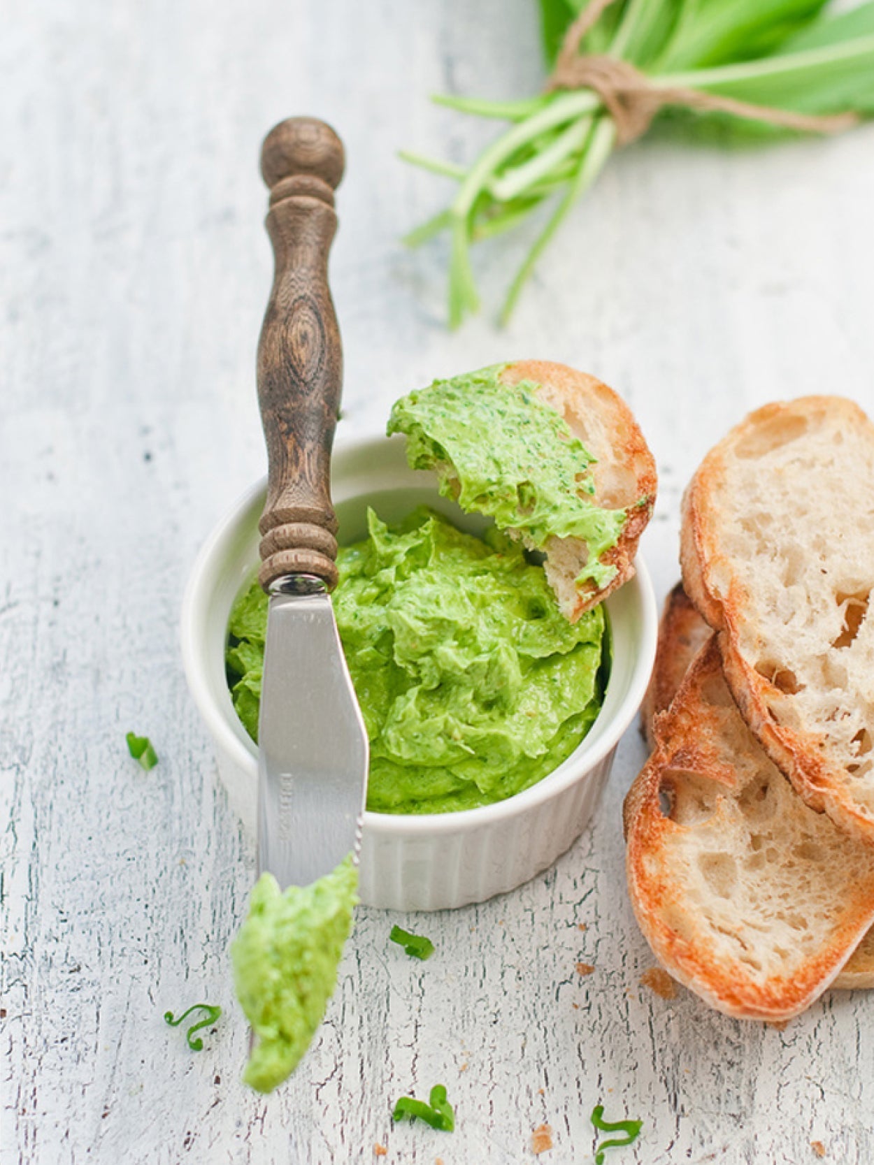 Cremige grüne Bärlauchbutter in einer weißen Schale mit rustikalem Holzmesser und frischem Baguette auf hellem Holztisch – frische Bärlauchblätter im Hintergrund.