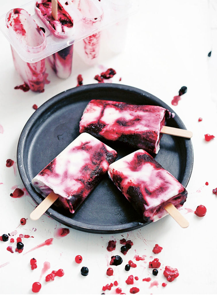 Drei Blaubeer-Joghurt-Eis am Stiel auf einem dunklen Teller, daneben Beeren und Eisformen auf hellem Untergrund.