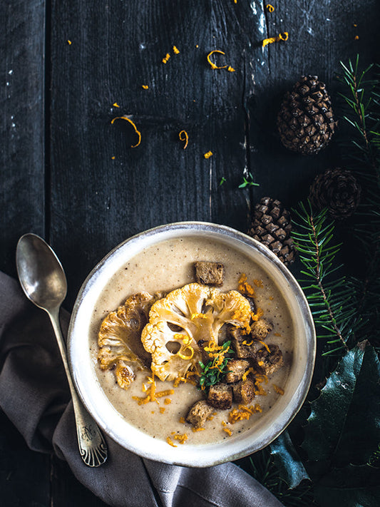 Cremige Blumenkohlsuppe in einer Schale, getoppt mit goldbraun gerösteten Blumenkohlscheiben, knusprigen Zimt-Croûtons und Orangen-Petersilien-Gremolata, dekorativ angerichtet auf dunklem rustikalem Holz mit Tannenzweigen und Zapfen.