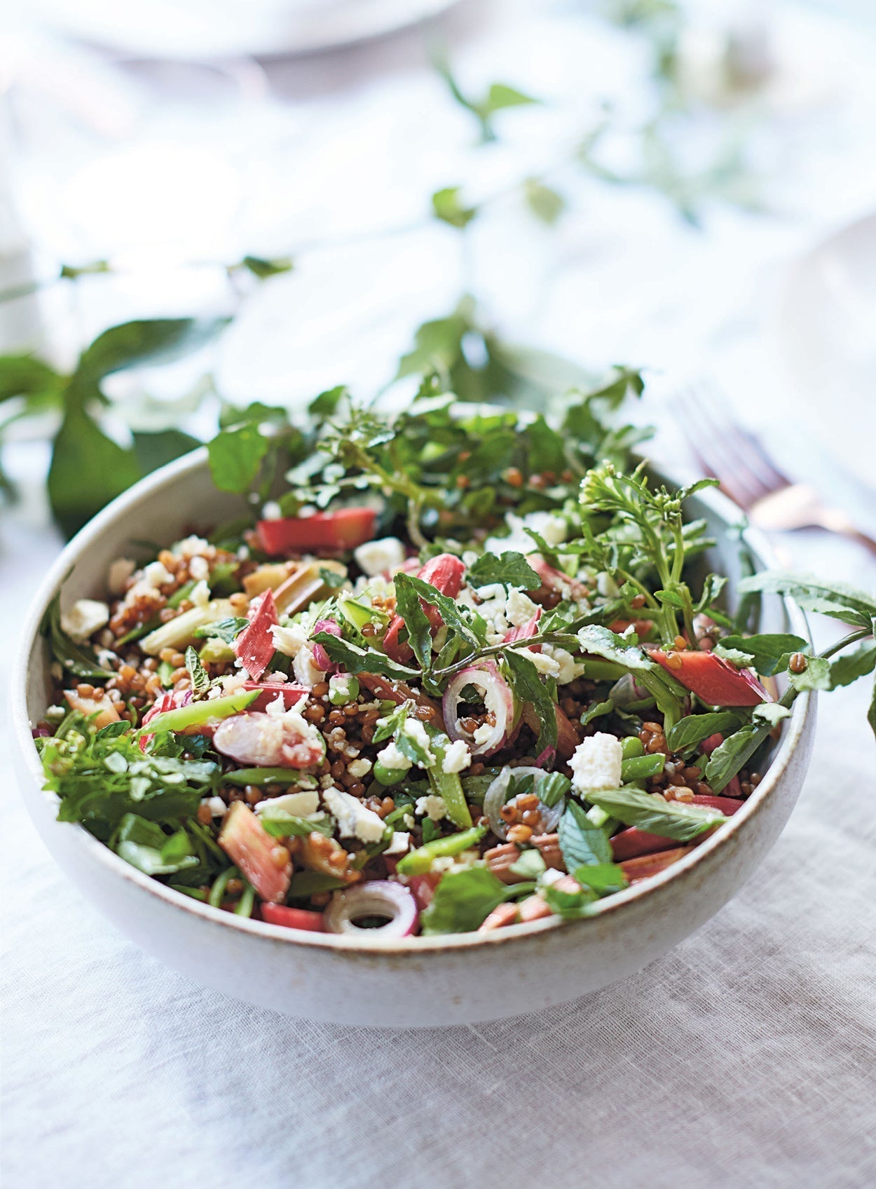 Frühlingshafte Weizen-Bowl mit eingelegtem Rhabarber, frischen Kräutern, Zuckerschoten, Radieschen und zerbröseltem Ziegenkäse in einer Keramikschüssel auf hellem Leinentuch