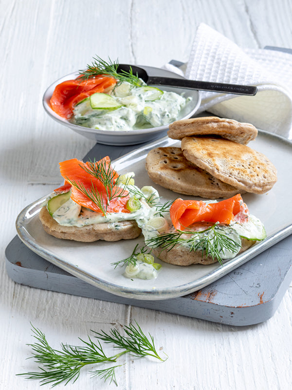 Herzhafte Buchweizen-Pancakes mit Gurken-Dill-Crème und Graved Lachs auf einem hellen Servierteller, garniert mit frischem Dill – sommerlich leichtes Gericht auf weißem Holztisch.