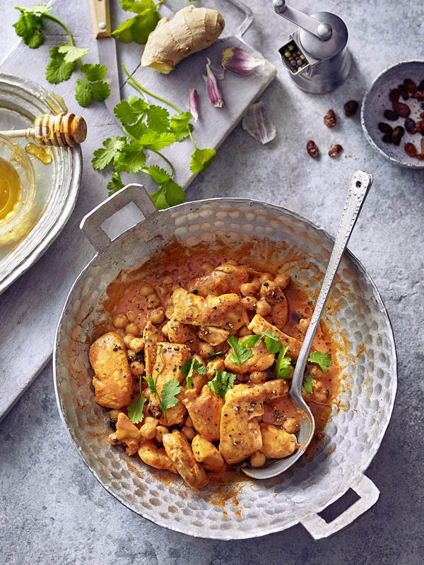 Eine Pfanne mit cremigem Butter-Chicken in Tomaten-Sahne-Sauce, angerichtet mit Kichererbsen und frischem Koriander; daneben Ingwer, Kräuter und Gewürze.